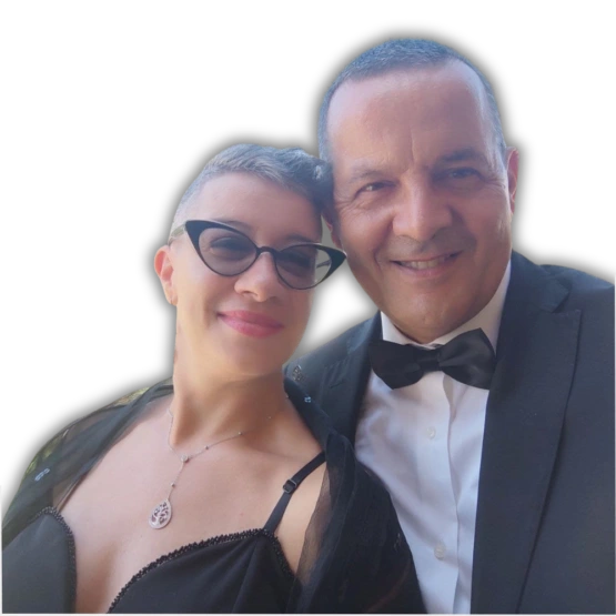 “Due persone, un uomo ed una donna, sorridenti posano insieme per una fotografia. Indossano abiti eleganti da sera; la ragazza indossa un abito scuro con accessori e occhiali, il ragazzo una giacca formale con camicia e papillon. Lo sfondo è neutro e scuro.”